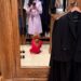 A woman in a lavender coat stands in front of a mirror, while a child in a red outfit sits on the floor.