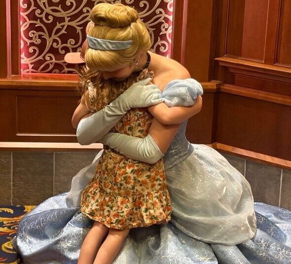 An individual dressed in a Cinderella costume is embracing another person, their faces obscured by the angle of the image. Both