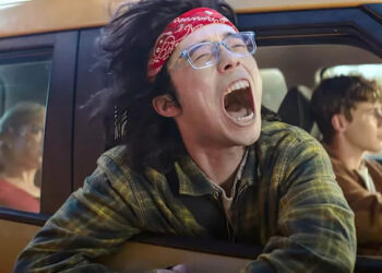 A group of teens ride in a yellow car, with one kid screaming with his head out the window.