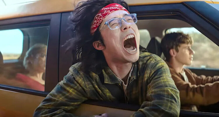 A group of teens ride in a yellow car, with one kid screaming with his head out the window.