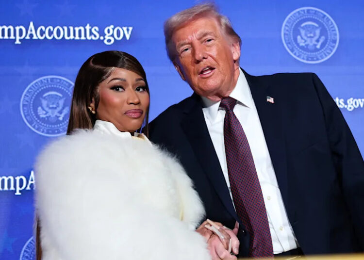 Nicki Minaj in a white fur coat and Donald Trump in a suit stand together at an event with a blue backdrop.
