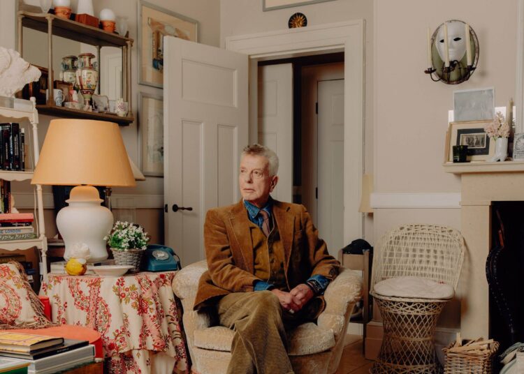 Nicky Haslam seated in the sitting room of his Cotswolds home wearing a brown jacket and trousers