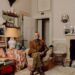 Nicky Haslam seated in the sitting room of his Cotswolds home wearing a brown jacket and trousers