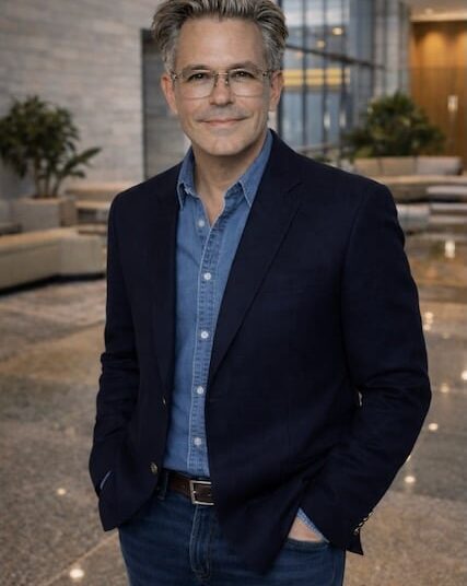 A person with glasses wearing a denim shirt and jacket.