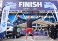 Singer Olly Murs crosses the finish line with his hands in the air wearing orange shorts and a black top