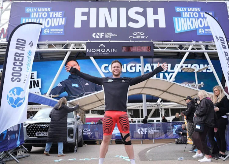 Singer Olly Murs crosses the finish line with his hands in the air wearing orange shorts and a black top