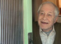 An older Rothwell wearing a shirt and cardigan and opening a door in character during a scene from Doctors.