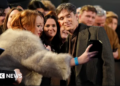 Cillian Murphy, wearing a dark-coloured suit and coat, is posing for a selfie with a woman wearing a fluffy coat. she is leaning over a barrier, and there are a large number of people alongside and behind her.