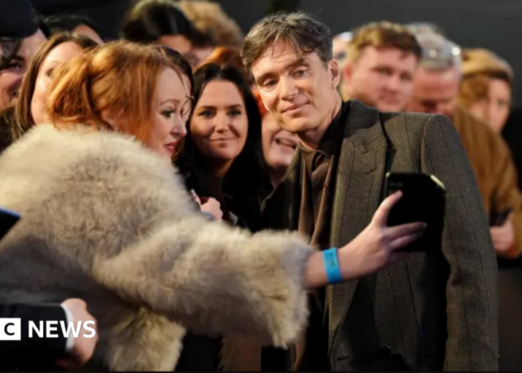 Cillian Murphy, wearing a dark-coloured suit and coat, is posing for a selfie with a woman wearing a fluffy coat. she is leaning over a barrier, and there are a large number of people alongside and behind her.