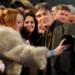 Cillian Murphy, wearing a dark-coloured suit and coat, is posing for a selfie with a woman wearing a fluffy coat. she is leaning over a barrier, and there are a large number of people alongside and behind her.