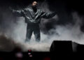 Ye stands on stage with arms outstretched, surrounded by smoke, wearing a dark jacket and pants.