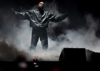 Ye stands on stage with arms outstretched, surrounded by smoke, wearing a dark jacket and pants.