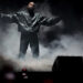 Ye stands on stage with arms outstretched, surrounded by smoke, wearing a dark jacket and pants.