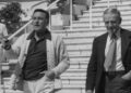 A black and white image of Gene Kelly and Fred Astaire walking down some steps in Cannes. Kelly is holding a pair of sunglasses and is wearing a dark polo shirt with a thick knit cardigan. Astaire is wearing a dark suit and tie. Both men have one hand in their pocket.