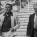 A black and white image of Gene Kelly and Fred Astaire walking down some steps in Cannes. Kelly is holding a pair of sunglasses and is wearing a dark polo shirt with a thick knit cardigan. Astaire is wearing a dark suit and tie. Both men have one hand in their pocket.