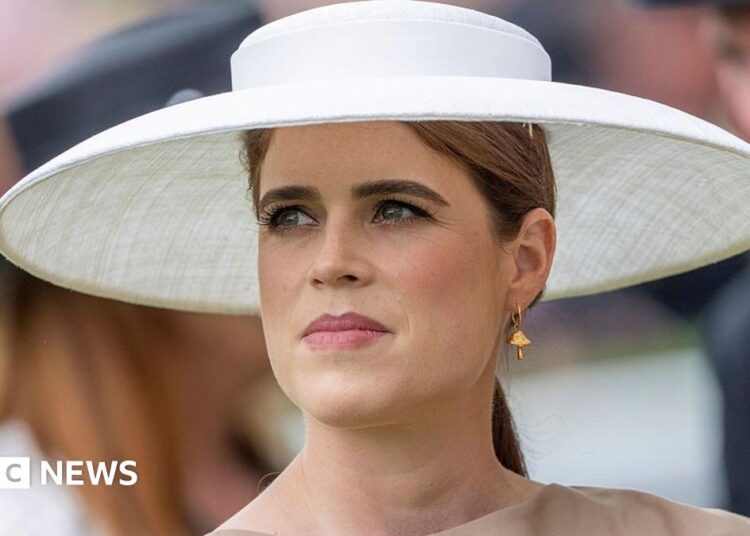 Princess Eugenie wearing a large wide-brimmed white hat, gold toadstool earring and brown dress