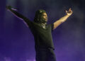Ken Carson on stage with arms outstretched, holding a microphone, wearing a black outfit and cap, against a purple-lit background.