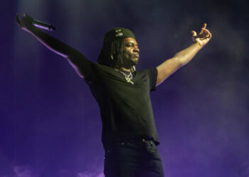 Ken Carson on stage with arms outstretched, holding a microphone, wearing a black outfit and cap, against a purple-lit background.
