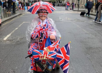 A man riding a mobility scooter covered in Union flags. He has a small umbrella attached to his hat, which also has a Union flag design.