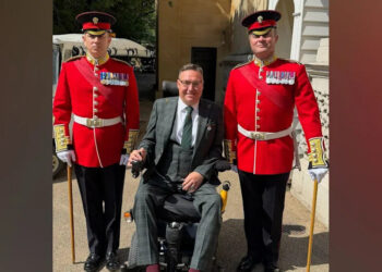 A man, wearing a grey suit, is sitting in a wheelchair. There are two guards, in red coats, standing beside him.