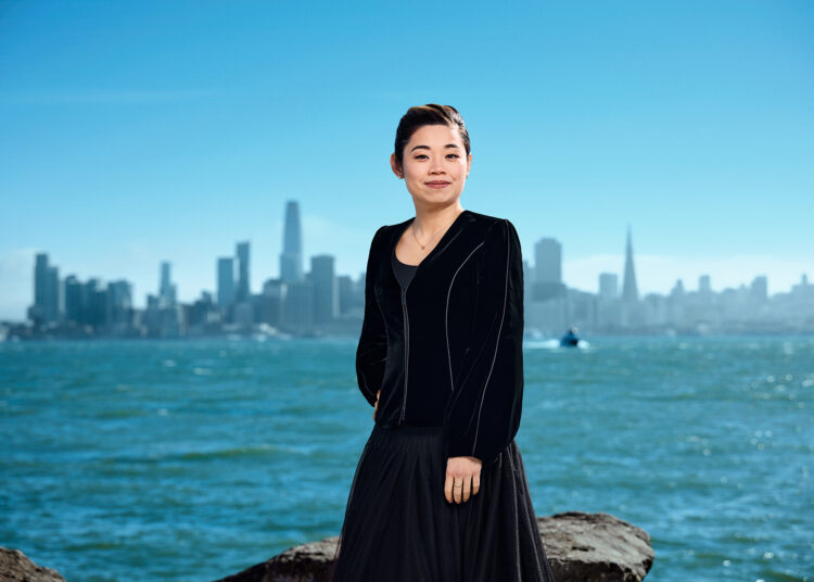 A young smiling Asian woman stands before a body of water with the San Francisco skyline in the background