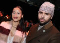 Chris Brown and Jada Wallace posing together at an event. Brown wears a beanie and coat, and Wallace wears a striped scarf.