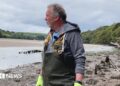 A person wearing red protective gloves uses a handheld power tool to grind or cut through a metal frame covered in seaweed. Bright sparks fly from the contact point as the work is carried out on a muddy tidal shoreline with shallow water in the background.