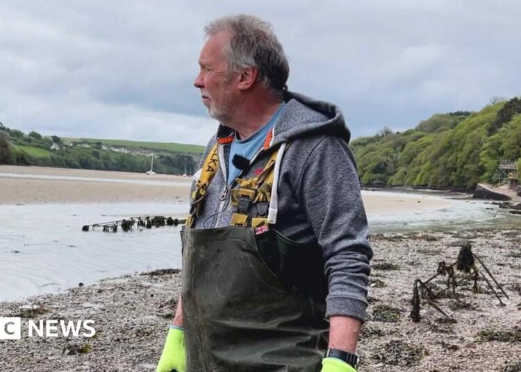 A person wearing red protective gloves uses a handheld power tool to grind or cut through a metal frame covered in seaweed. Bright sparks fly from the contact point as the work is carried out on a muddy tidal shoreline with shallow water in the background.