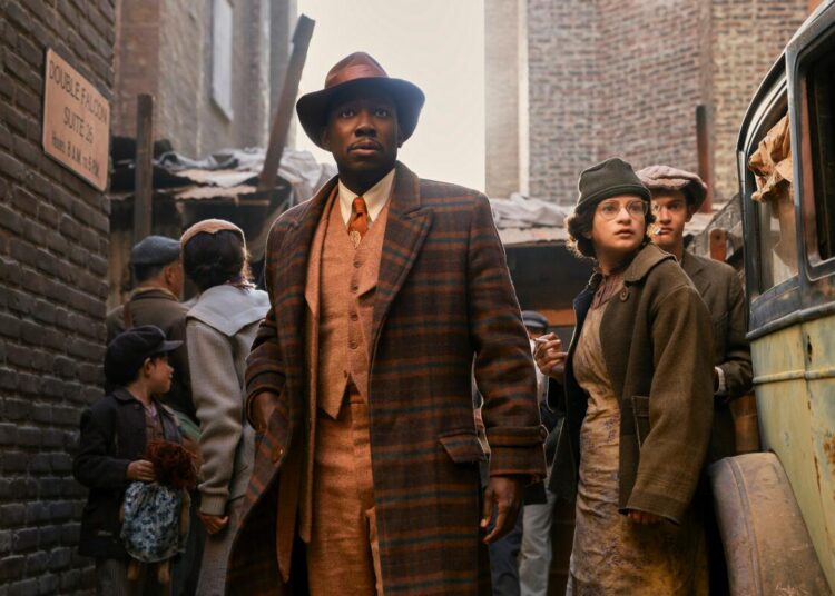 A man in a plaid coat, orange-hued suit and brown hat stands in an alleyway crowded with people.