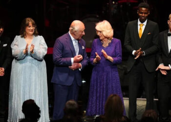 The monarch is dressed in what appears to be a navy blue suit, stood next to his wife, who is wearing a pattered purple dress. They are flanked by award winners who are in dinner jackets