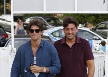 Two individuals, dressed in casual summer attire, are strolling along a coastal pathway. The person on the left is wearing a light blue shirt and patterned shorts, while the person on the right is donning a maroon shirt and similar patterned shorts. They both appear to be engaged in casual conversation, with the surrounding environment suggesting a beachside location.