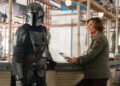 A man in a mask stands next to a woman in a green coat.