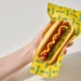 A person holding a Spam hot dog with ketchup, mustard and relish on a yellow wrapper