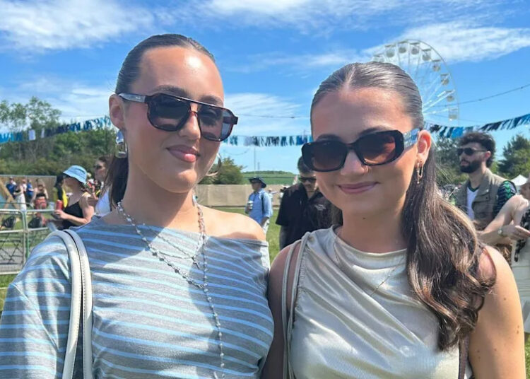 Two young women in sunglasses smile at the camera on a music festival site.