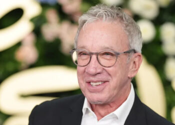 Tim Allen smiling, wearing glasses and a suit, in front of a floral backdrop.