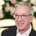 Tim Allen smiling, wearing glasses and a suit, in front of a floral backdrop.