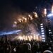 A massive outdoor festival crowd at night with arms raised toward a large stage structure topped with flames and confetti cannons firing silver streamers into the air, with bright white and amber stage lights cutting through the smoke and darkness above the packed crowd.