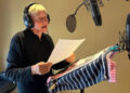 A person with short gray hair wearing headphones stands at a microphone, reading from a script on a music stand covered with striped cloths in a recording studio.