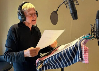 A person with short gray hair wearing headphones stands at a microphone, reading from a script on a music stand covered with striped cloths in a recording studio.
