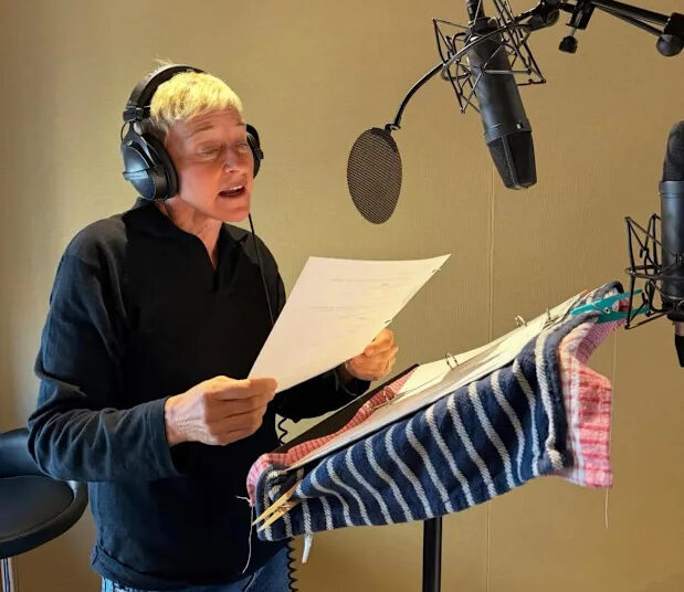 A person with short gray hair wearing headphones stands at a microphone, reading from a script on a music stand covered with striped cloths in a recording studio.