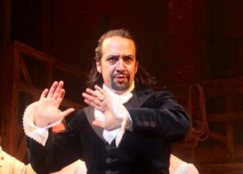 A man in historical clothing with lace cuffs gestures with both hands raised and a serious expression, standing on a dimly lit stage with wooden beams and ropes in the background.