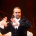 A man in historical clothing with lace cuffs gestures with both hands raised and a serious expression, standing on a dimly lit stage with wooden beams and ropes in the background.