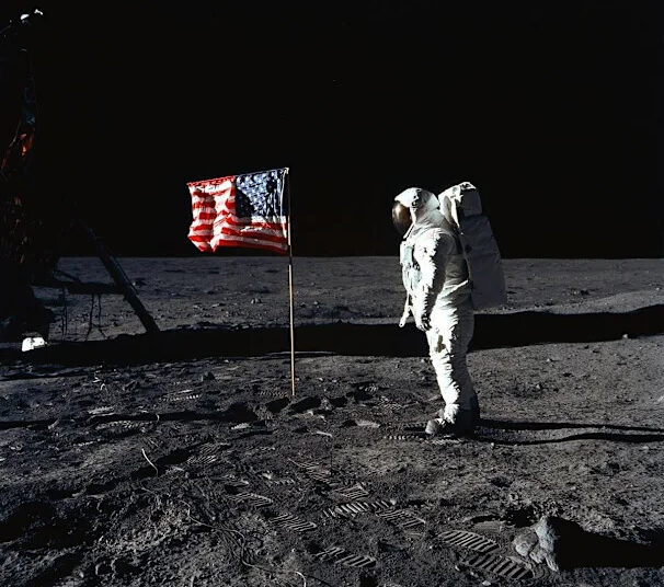 An astronaut in a spacesuit stands on the Moon next to an American flag, with lunar lander parts and footprints visible on the gray, dusty surface against a black sky.