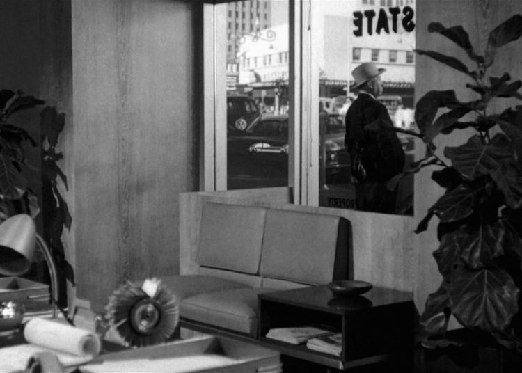 A black-and-white image of a modern office with large plants, a desk with various objects, a couch, and a window. Outside the window, a person in a hat stands on the sidewalk with buildings and cars visible across the street.