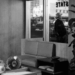 A black-and-white image of a modern office with large plants, a desk with various objects, a couch, and a window. Outside the window, a person in a hat stands on the sidewalk with buildings and cars visible across the street.