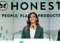 A woman in a green blazer stands at a podium, speaking. Behind her is a large sign that reads "HONEST" and "People. Planet. Products." She appears confident and professional.