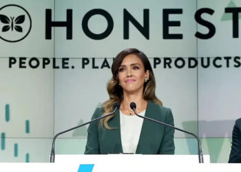 A woman in a green blazer stands at a podium, speaking. Behind her is a large sign that reads "HONEST" and "People. Planet. Products." She appears confident and professional.