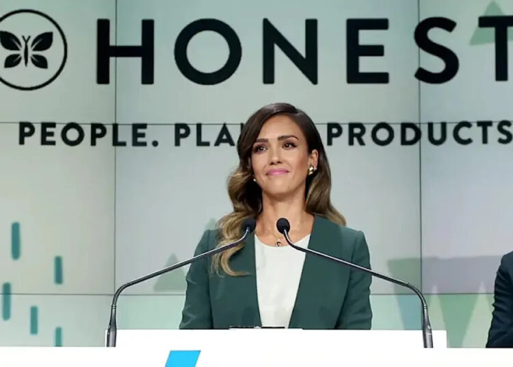 A woman in a green blazer stands at a podium, speaking. Behind her is a large sign that reads "HONEST" and "People. Planet. Products." She appears confident and professional.
