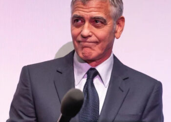 A man in a dark suit and tie holds a microphone, looking to the side with a slight, closed-mouth smile against a light-colored background.