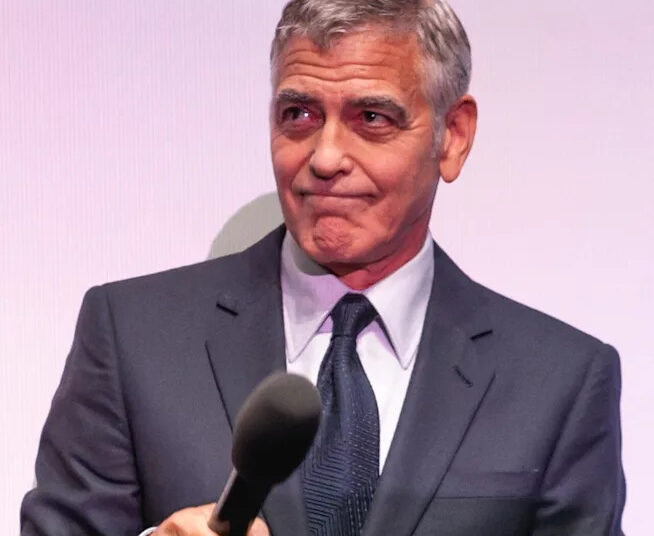 A man in a dark suit and tie holds a microphone, looking to the side with a slight, closed-mouth smile against a light-colored background.